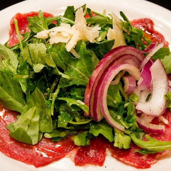 Thinly sliced beef carpaccio topped with arugula, red onions, shaved Parmesan, and olive oil on a white plate.