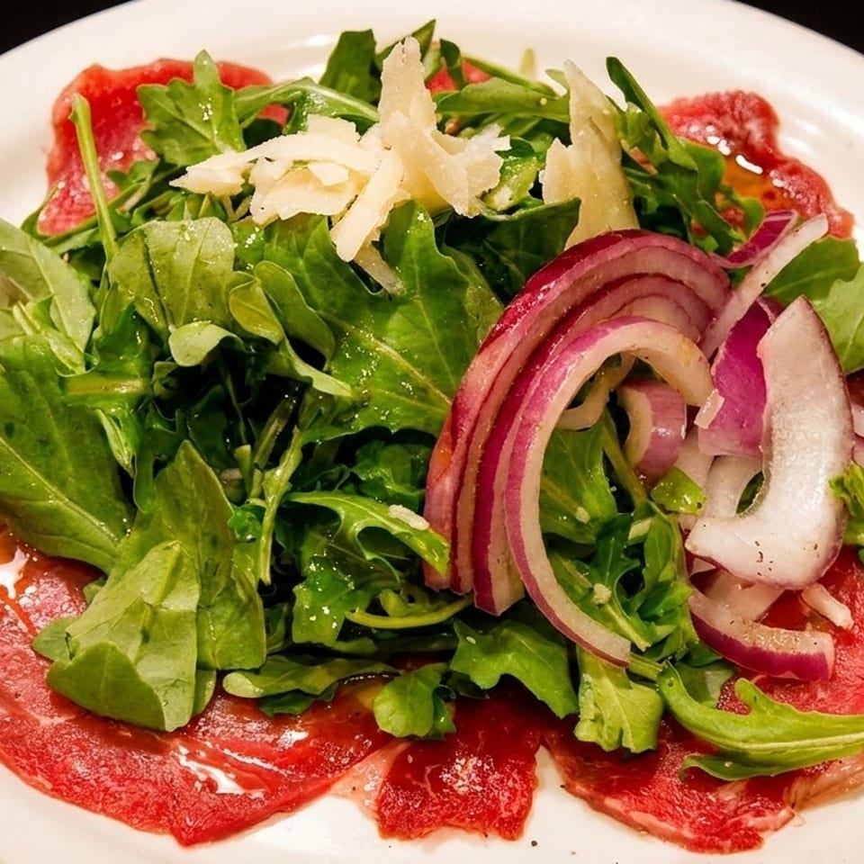Thinly sliced beef carpaccio topped with arugula, red onions, shaved Parmesan, and olive oil on a white plate.