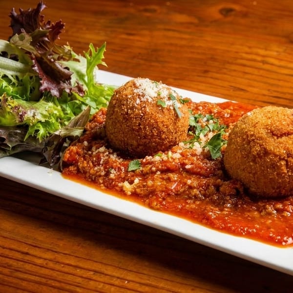 Two golden-brown arancini balls on a bed of marinara sauce, with a side salad on a white plate.