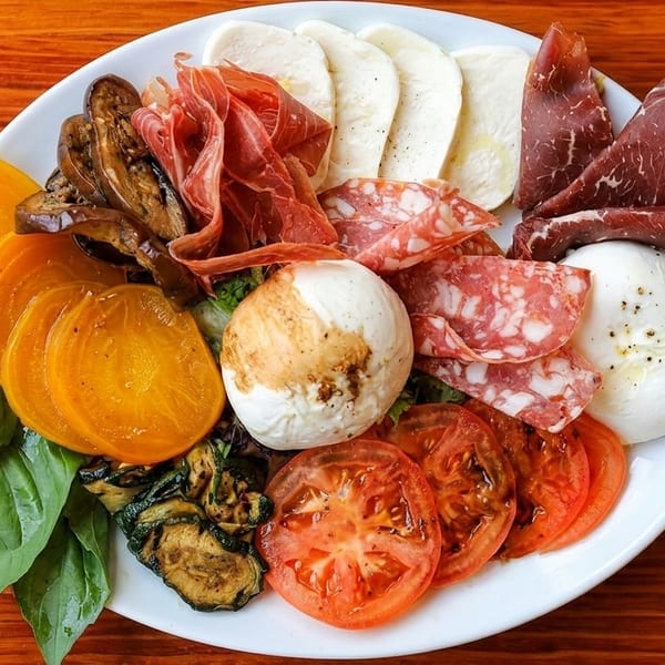 A white plate filled with charcuterie, cheeses, grilled vegetables, and fresh basil on a wooden table.